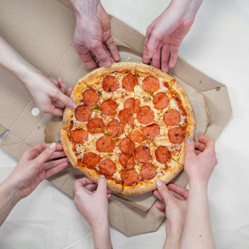 Seven different hands grab a slice of a salami pizza cut in eight different pieces.